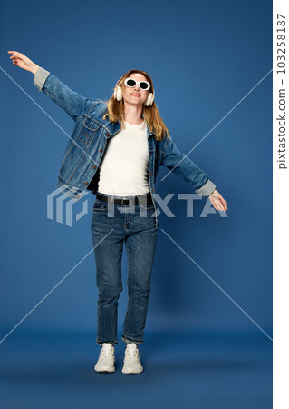 Full-length portrait of young girl in jeans outfit listening to music in headphones, smiling against blue studio background Full-length portrait of young girl in jeans outfit listening to music in headphones, smiling against blue studio background 103258187