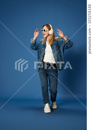 Full-length portrait of young girl in jeans outfit listening to music in headphones, smiling against blue studio background Full-length portrait of young girl in jeans outfit listening to music in headphones, smiling against blue studio background 103258188