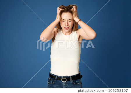 Portrait of young girl holding head with hands, suffering from stress and depression against blue studio background Portrait of young girl holding head with hands, suffering from stress and depression against blue studio background 103258197
