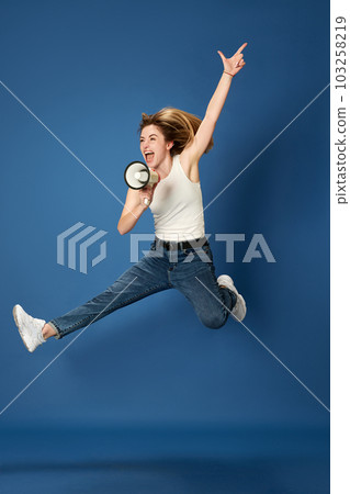 Full-length portrait of young woman in casual clothes posing, jumping and shouting in megaphone against blue studio background. News 103258219