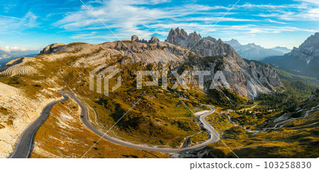 Road winds at foot of rocky mountain in Tre Cime di Lavaredo. Scenic mountain ranges under blue sky with white clouds at sunset aerial view Road winds at foot of rocky mountain in Tre Cime di Lavaredo. Scenic mountain ranges under blue sky with white clouds at sunset aerial view 103258830