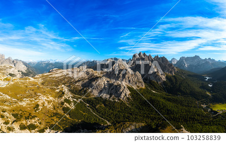 Rocky mountain peaks under blue sky with white clouds. Covered with grey sand deep canyons. Mountains near theThree Peaks of Lavaredo at sunset aerial view Rocky mountain peaks under blue sky with white clouds. Covered with grey sand deep canyons. Mountains near theThree Peaks of Lavaredo at sunset aerial view 103258839
