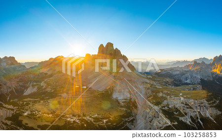 Sun rises over mountain range. Rocky mountains with sand-covered foothills and green valley. Three Peaks of Lavaredo at sunrise aerial view in back lit Sun rises over mountain range. Rocky mountains with sand-covered foothills and green valley. Three Peaks of Lavaredo at sunrise aerial view in back lit 103258853