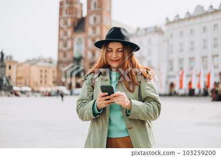 Beautiful stylish woman in hat walking on Market Square in Krakow on autumn day and holding mobile phone. Urban lifestyle concept. Check social networks, send sms or book hotel Beautiful stylish woman in hat walking on Market Square in Krakow on autumn day and holding mobile phone. Urban lifestyle concept. Check social networks, send sms or book hotel 103259200