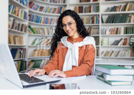 Young hispanic woman studying in academic university library, female student smiling and looking at camera while sitting at laptop, woman with curly hair. 103260864