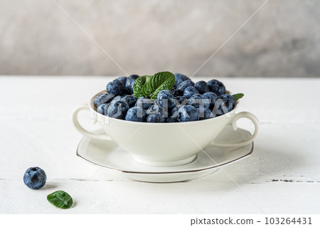 Fresh blueberries in a white teacup on the white wooden table with grey concrete background, closeup, free space for text 103264431
