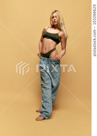 Young beautiful woman with slim body, posing in giant, oversized jeans against yellow studio background. Weight loss, sport and diet Young beautiful woman with slim body, posing in giant, oversized jeans against yellow studio background. Weight loss, sport and diet 103266629