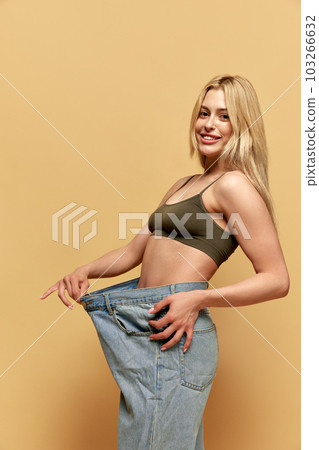 Young beautiful woman with slim body, posing in giant, oversized jeans against yellow studio background. Weight loss, sport and diet Young beautiful woman with slim body, posing in giant, oversized jeans against yellow studio background. Weight loss, sport and diet 103266632