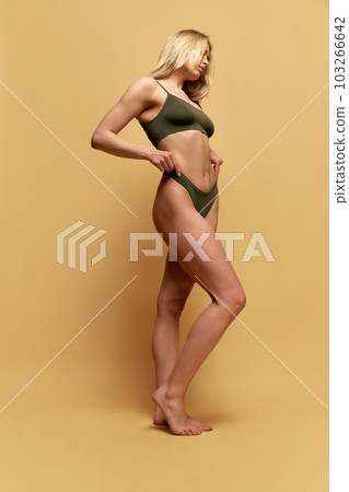 Full-length image of young blonde woman with slim, fit, muscular body posing in green underwear a against yellow studio background 103266642