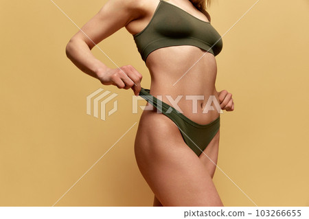 Cropped image of slim, fit female body, breast, belly, legs in green underwear posing against yellow studio background Cropped image of slim, fit female body, breast, belly, legs in green underwear posing against yellow studio background 103266655