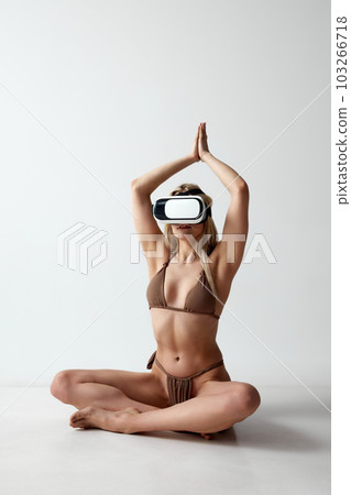 Young blonde girl with slim, tanned, fit body in swimsuit sitting in vr glassess against grey studio background. Online training Young blonde girl with slim, tanned, fit body in swimsuit sitting in vr glassess against grey studio background. Online training 103266718