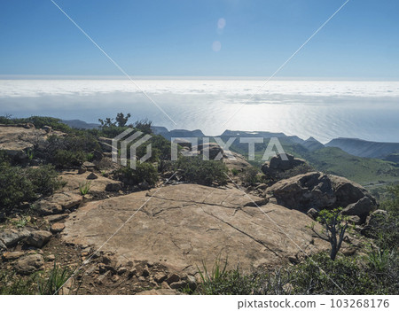 View of top of table mountain Fortaleza. Flat rock and stones, green hills and cliffs. Ocean and white clouds above. Blue sky background. La Gomera, Canary Islands, Spain. 103268176