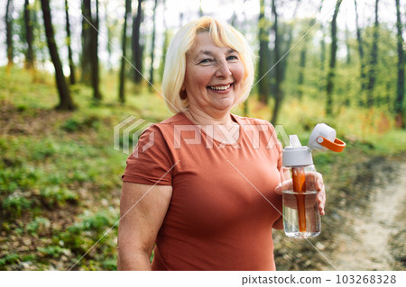 Image of senior 60s woman using earphones and drinking water while working out near road bridge in morning in a park Image of senior 60s woman using earphones and drinking water while working out near road bridge in morning in a park 103268328