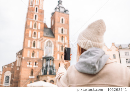 Young smile woman tourist In a stylish coat takes selfie on mobile phone on the market square in Krakow in Poland Young smile woman tourist In a stylish coat takes selfie on mobile phone on the market square in Krakow in Poland 103268341