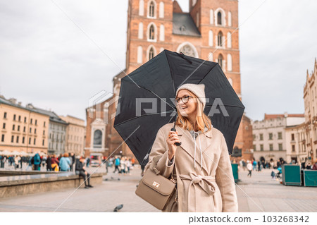 Young blonde woman smiles under a black umbrella breathing in the street of and old town Krakow in a rainy day. High quality photo. Natural beauty Young blonde woman smiles under a black umbrella breathing in the street of and old town Krakow in a rainy day. High quality photo. Natural beauty 103268342