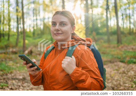 Side view of young female traveler with backpack and in warm wearing standing in the spring forest looking at phone screen. Women on hiking trail. Wooden path in forest. National Park in Poland 103268355