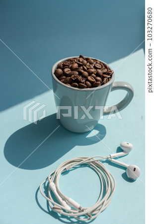 Black coffee beans in blue cup and white headphones on blue background with sunlight shadow top view. Morning drink concept. Cafe and breakfast background. Roasted coffee mug. Aroma drink. Black coffee beans in blue cup and white headphones on blue background with sunlight shadow top view. Morning drink concept. Cafe and breakfast background. Roasted coffee mug. Aroma drink. 103270680