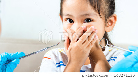 Dental kid examination. Doctor examines oral cavity of child uses mouth mirror to check teeth cavity but little girl cover mouth no need to checking she afraid, scared and closes his mouth with hands 103271139