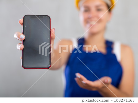 Smiling female worker in work clothes and hardhat demonstrating screen of her phone Smiling female worker in work clothes and hardhat demonstrating screen of her phone 103271163