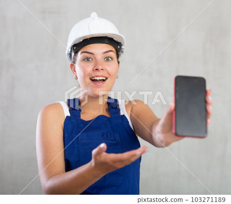 Smiling female worker in work clothes and hardhat demonstrating screen of her phone 103271189