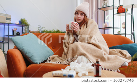 Sick ill woman suffering from cold drinking hot tea sitting on couch sneezes wipes snot into napkin Sick ill woman suffering from cold drinking hot tea sitting on couch sneezes wipes snot into napkin 103273746