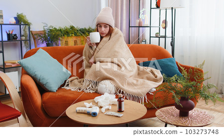Sick ill woman suffering from cold or allergy sitting on home sofa sneezes wipes snot into napkin Sick ill woman suffering from cold or allergy sitting on home sofa sneezes wipes snot into napkin 103273748
