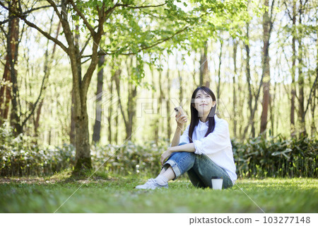 一位 30 多歲的日本女性在森林裡放鬆時操作智能手機的初夏形象 103277148