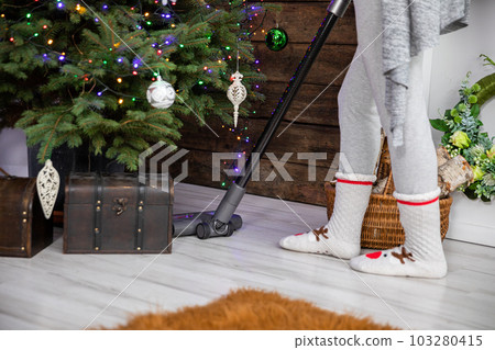 In the living room, a girl cleans thoroughly with a vacuum cleaner under the Christmas tree. In the living room, a girl cleans thoroughly with a vacuum cleaner under the Christmas tree. 103280415