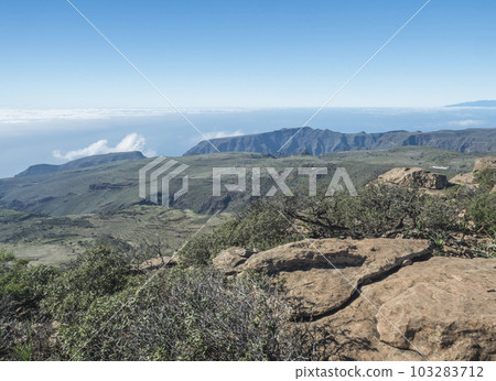 Scenic view from top of the table mountain Fortaleza. Rock, green hills and plains, white clouds and El Hiero island above, blue sky. La Gomera, Canary Islands, Spain. 103283712
