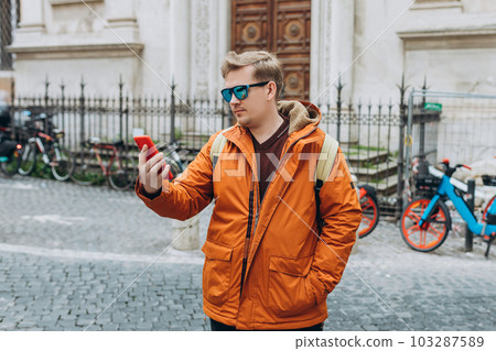 Cheerful young guy walking on city street with modern smartphone in hands. Handsome man in sunglasses reading some good and smiling. Technology for lifestyles. 103287589