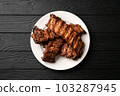 Freshly cooked pork ribs on a white round plate on a black wooden table in the background. Close up 103287945