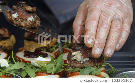 A chef preparing mini burgers for his customer, puts grilled meat on buns with vegetables. 103295367