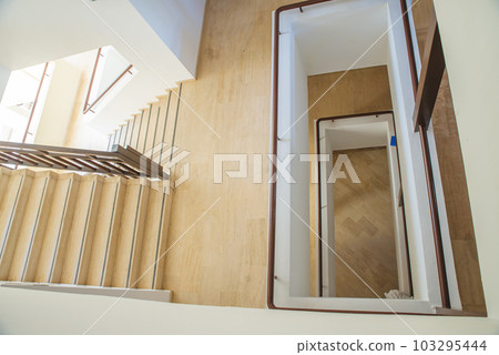 Hotel staircase with slightly curved architecture, windows and historic metal railing. Stairs are tiled and the handrail is supplemented with wooden handrail. There is also wood on walls and paneling. 103295444