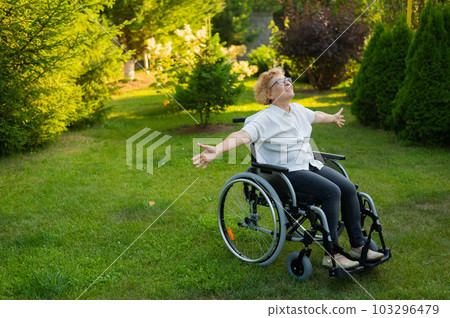 Happy old woman spread her arms to the sides while sitting in a wheelchair on a walk outdoors. Happy old woman spread her arms to the sides while sitting in a wheelchair on a walk outdoors. 103296479