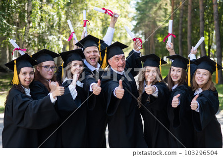 A group of graduates in robes give a thumbs up outdoors. Elderly student. 103296504