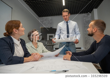 A caucasian man stands and holds a drawing, three colleagues sit at a table and listen to him.  103296509