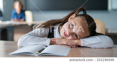 Little caucasian girl is bored at the lesson at school. The schoolgirl folded her head on the desk and the teacher sits in the background. Little caucasian girl is bored at the lesson at school. The schoolgirl folded her head on the desk and the teacher sits in the background. 103296896
