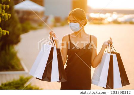 Fashionable woman wearing protective medical mask with shopping bags at sunset. Covid- 2019. Fashionable woman wearing protective medical mask with shopping bags at sunset. Covid- 2019. 103298312