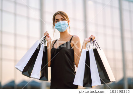 Beautiful woman in protective sterile medical mask with shopping bags near shopping center.Covid- 2019. 103298335