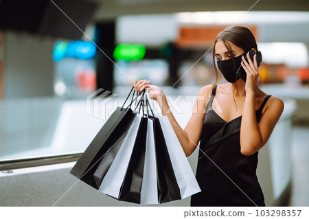 Young woman in protective black medical mask with shopping bags talking on a mobile phone in the mall. Young woman in protective black medical mask with shopping bags talking on a mobile phone in the mall. 103298357