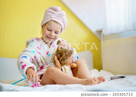 Cute smiling little girl child playing with her doll at home. Preschool child in bathrobe after taking bath brushing hairs of toy doll. Happy kid. Cute smiling little girl child playing with her doll at home. Preschool child in bathrobe after taking bath brushing hairs of toy doll. Happy kid. 103305713