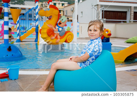 Little preschool girl sitting by outdoor swimming pool in hotel resort. Child learning to swim in outdoor pool, splashing with water, enjoying, laughing and having fun. Family vacations. Little preschool girl sitting by outdoor swimming pool in hotel resort. Child learning to swim in outdoor pool, splashing with water, enjoying, laughing and having fun. Family vacations. 103305875