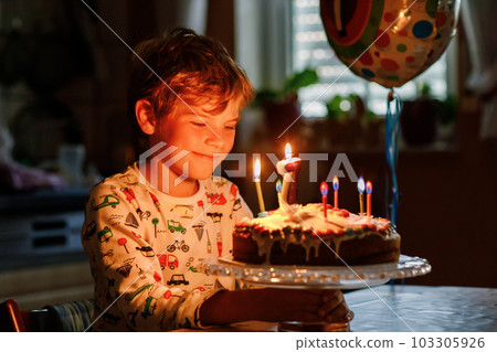 Adorable happy blond little kid boy celebrating his birthday. Child blowing seven candles on homemade baked cake, indoor. Birthday party for school children, family celebration of 7 years 103305926