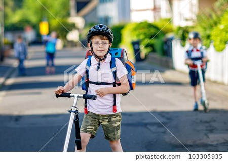 Two school kid boys in safety helmet riding with scooter in the city with backpack on sunny day. Happy children in colorful clothes biking on way to school. Safe way for kids outdoors to school 103305935