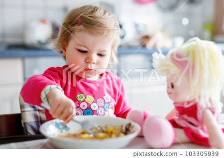 Adorable baby girl eating from fork vegetables and pasta. food, child, feeding and development concept. Cute toddler, daughter with spoon sitting in highchair and learning to eat by itself. 103305939
