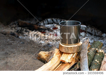 A metal mug for camping stands on firewood by the fire. Camping dishes, fire, evening. Copyspace 103308496