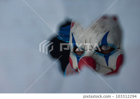 Creepy clown face peering through a hole in the partition. The face of a man wearing a creepy clown mask peering at his victim through a hole. A clown madman. 103312294