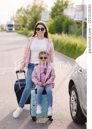 Adorable little girl sits on her kid suitcase and waiting for the trip with her mom. Two beautiful girls prepare to journey. A car is parked on the side 103313691