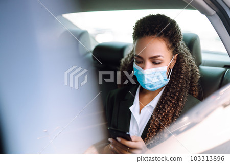 Woman in a sterile mask in taxi car on a back seat looking out of window checking her cell phone. 103313896