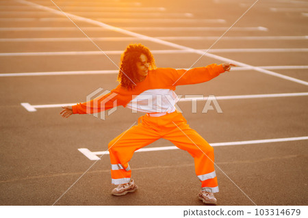 Young African American woman - dancer dancing in the street at sunset. Stylish woman with curly hair in an orange suit showing some moves. Young African American woman - dancer dancing in the street at sunset. Stylish woman with curly hair in an orange suit showing some moves. 103314679